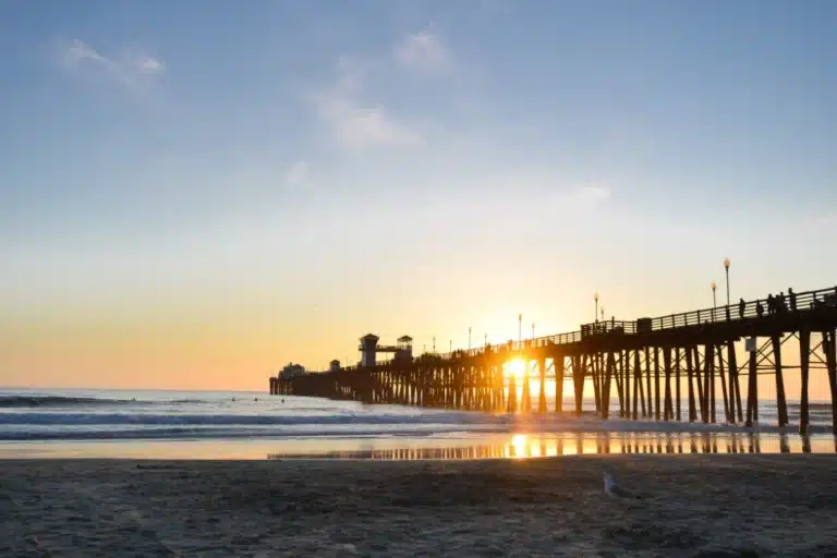 The sun setting behind Oceanside Pier, casting reflections on the wet sand and ocean waves, with silhouettes of people on the pier.