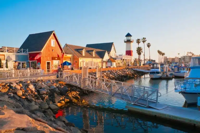 Colorful harbor village in Oceanside, California, featuring quaint shops, a striped lighthouse, and docked boats at sunset.