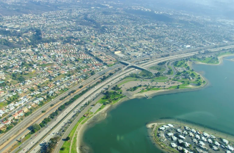 Aerial view of Mission Beach and surrounding neighborhoods with a coastal bay, highway interchange, dense housing, and waterfront park areas under a clear sky.