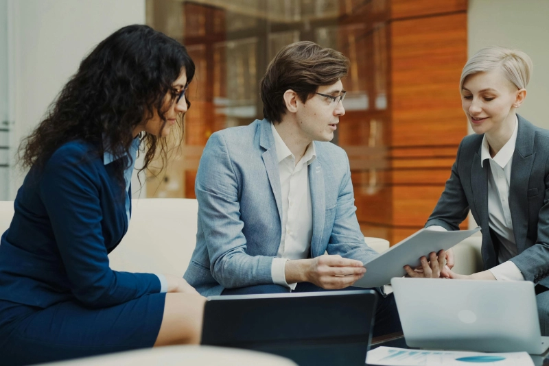 Three business professionals collaborate at a table, focused on a laptop during a meeting.