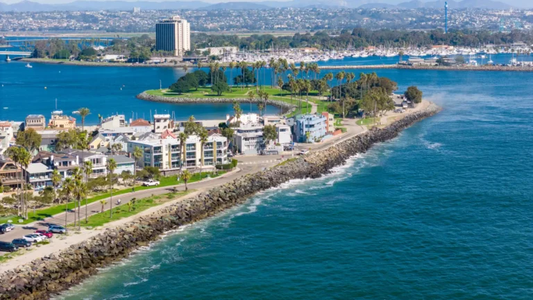 Aerial view of Mission Beach coastline with ocean waves, waterfront homes, a rocky shoreline, and a small island park extending into the bay.