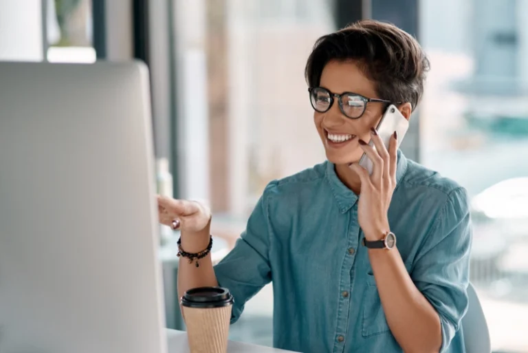 A woman wearing glasses speaks on the phone while seated at a desk, surrounded by office supplies.