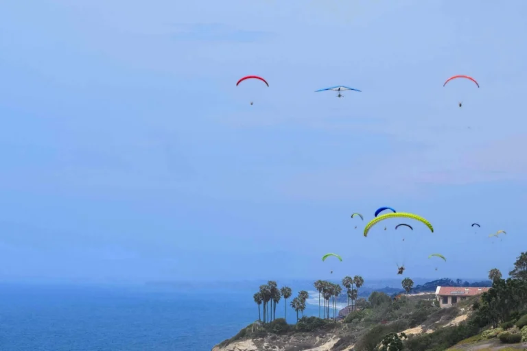 A diverse group of people joyfully flying colorful kites on a sunny hillside, surrounded by green grass and blue sky.